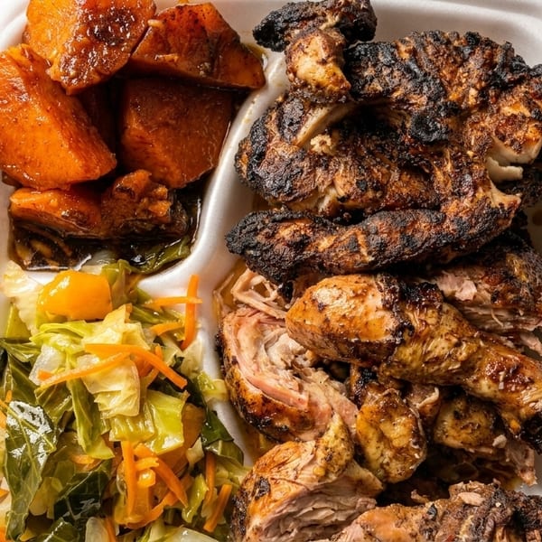 Takeout container with jerk chicken, candied yams, and collard greens on a wooden table.